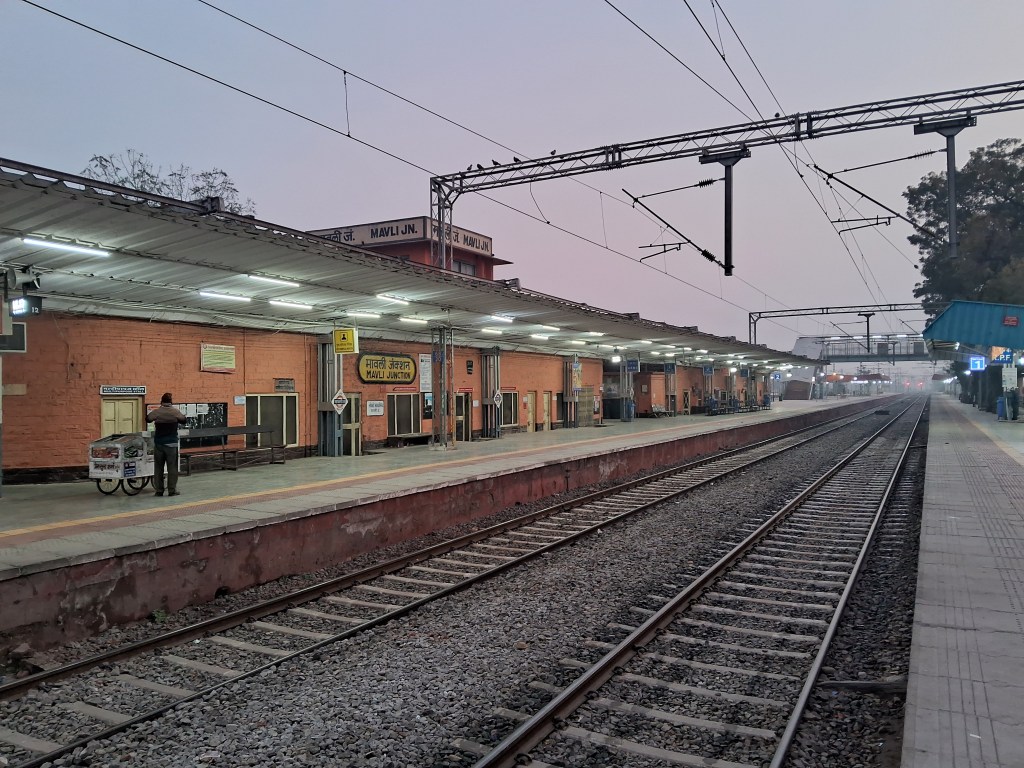 Mavli Junction Railway Station in the early morning, with empty platforms, silent tracks, and soft golden sunrise light creating a calm and peaceful atmosphere.