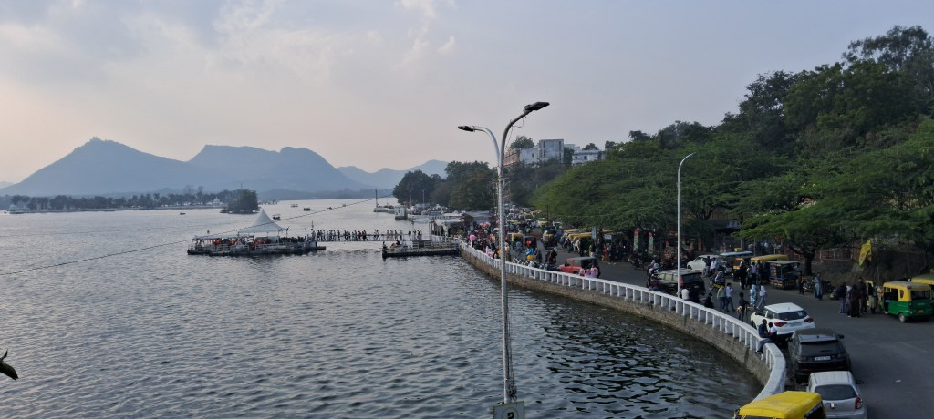 Serene view of Fateh Sagar Lake in Udaipur with a quiet driveway along its shore and the Aravalli hills rising in the background under a clear sky.