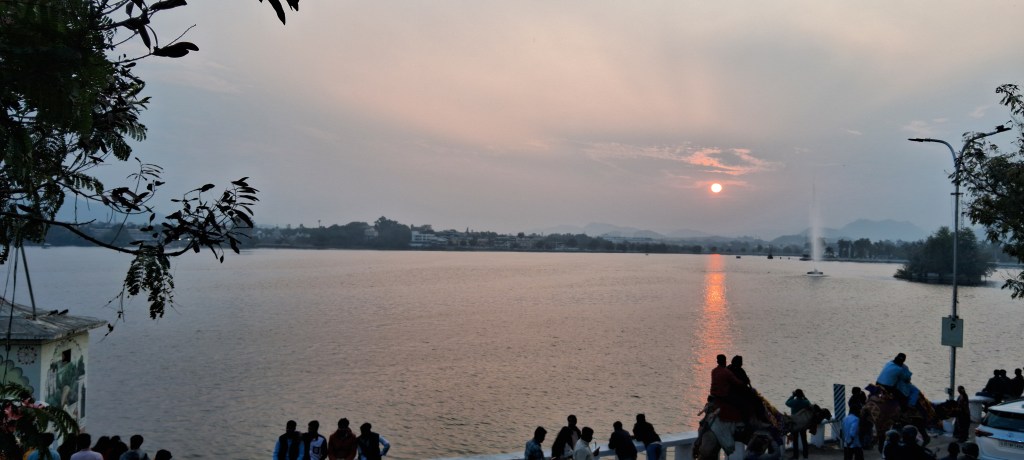 Scenic view of the saffron-tinted sun behind Fateh Sagar Lake in Udaipur just before sunset, with calm waters and silhouetted surroundings.
