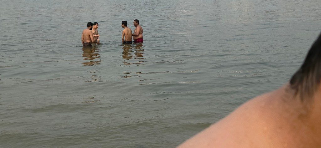 Devotees immersing in the holy Ganga River at Varanasi's ghats, with crowds wading through the flowing waters during a ritual bath and traditional attire billowing.