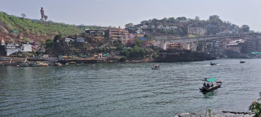 The calm flow of the Narmada River defines the beauty of Omkareshwar, with the distant presence of Adi Shankaracharya enriching the view.